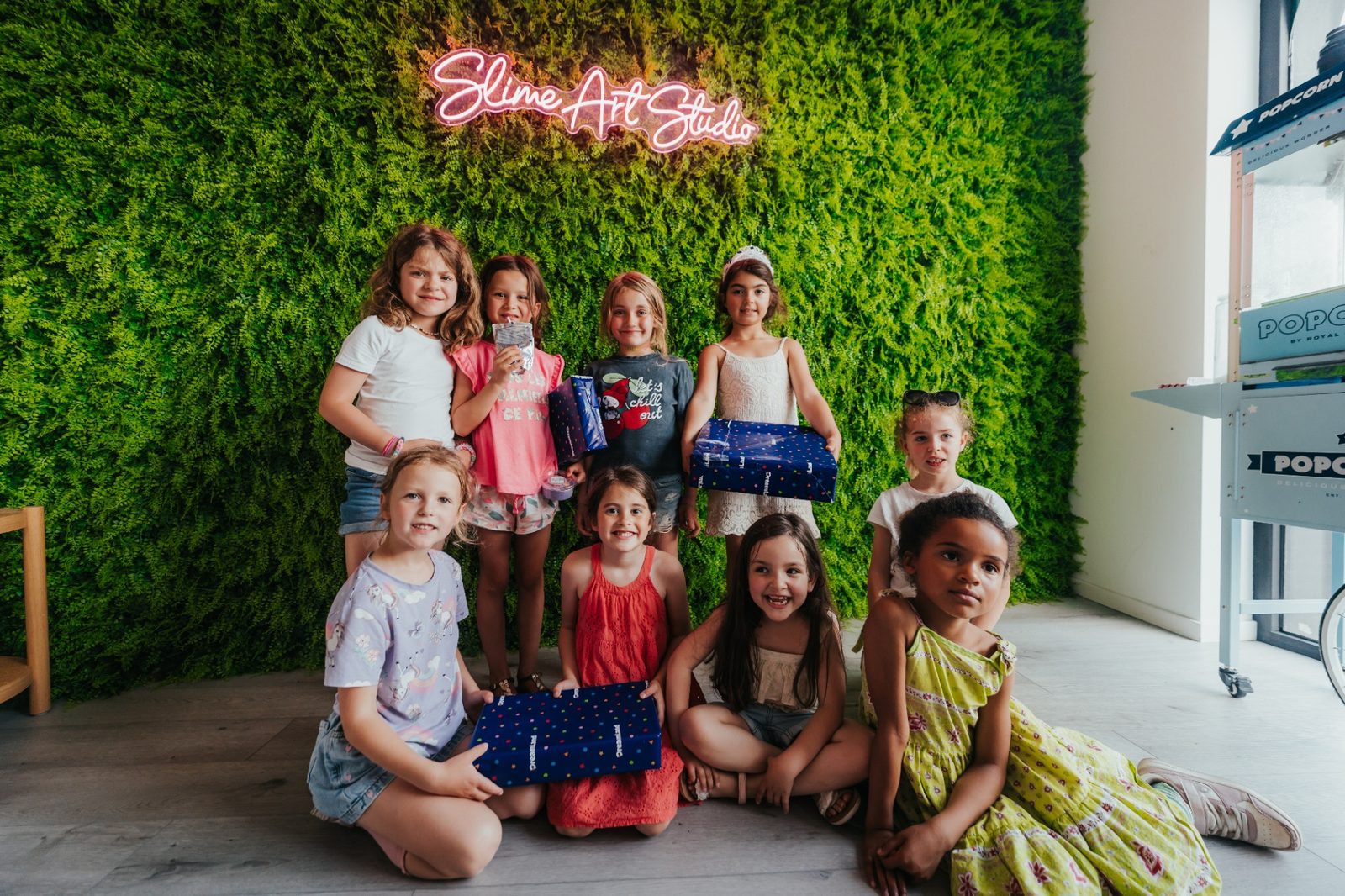 Slime Art Studio — groupe de neuf petites filles souriantes posant avec leurs pots de slime devant le mur végétal et l'enseigne néon Slime Art Studio, à côté de la machine à pop-corn