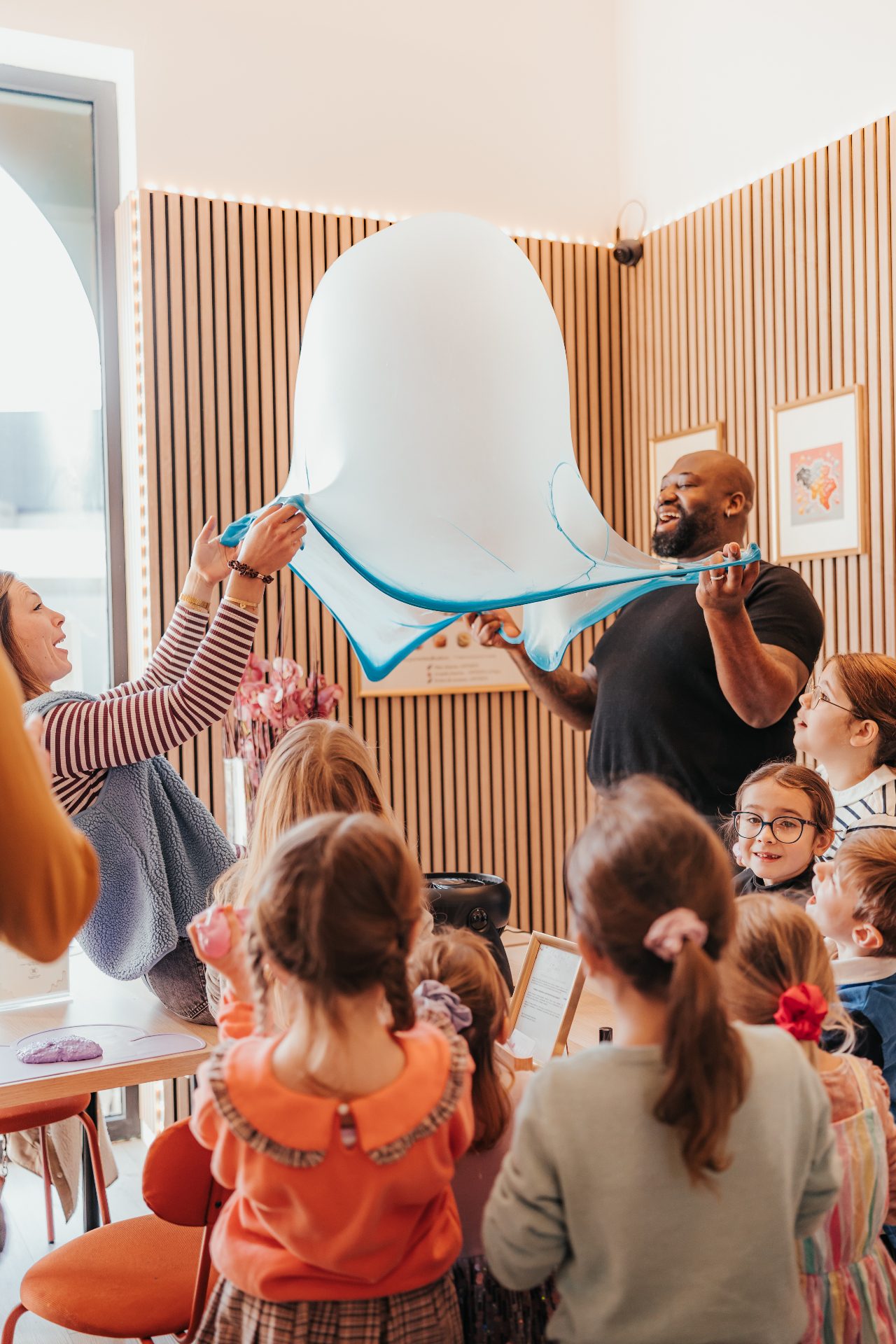 Atelier Slime Art Studio — animateurs souriants étirant un immense slime bleu et blanc devant un groupe d'enfants émerveillés dans une salle lumineuse aux murs en tasseaux de bois
