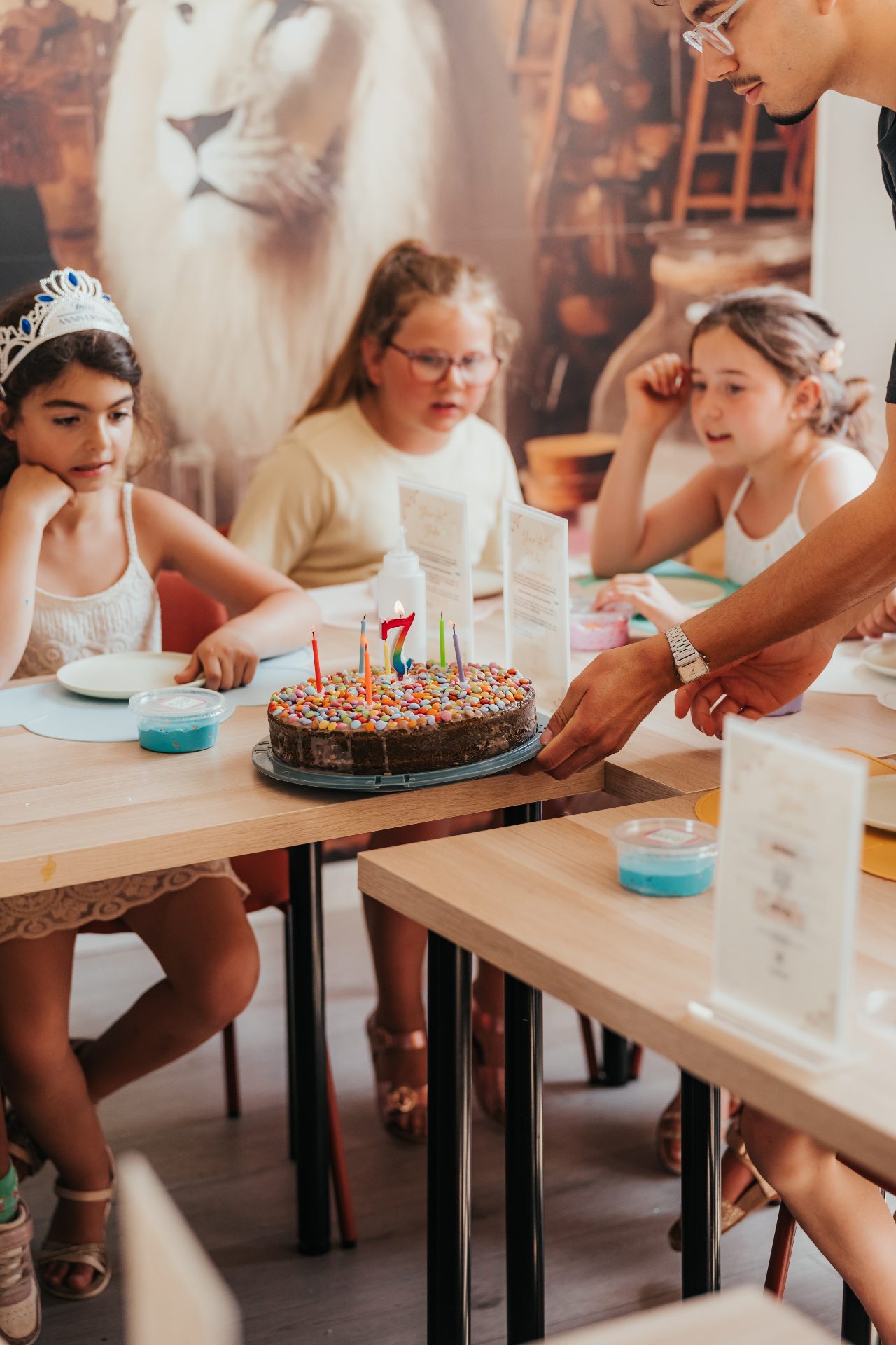 Anniversaire chez Slime Art Studio — animateur apportant un gâteau au chocolat aux Smarties avec bougie numéro 7 à trois petites filles attablées, dont une portant une couronne d'anniversaire