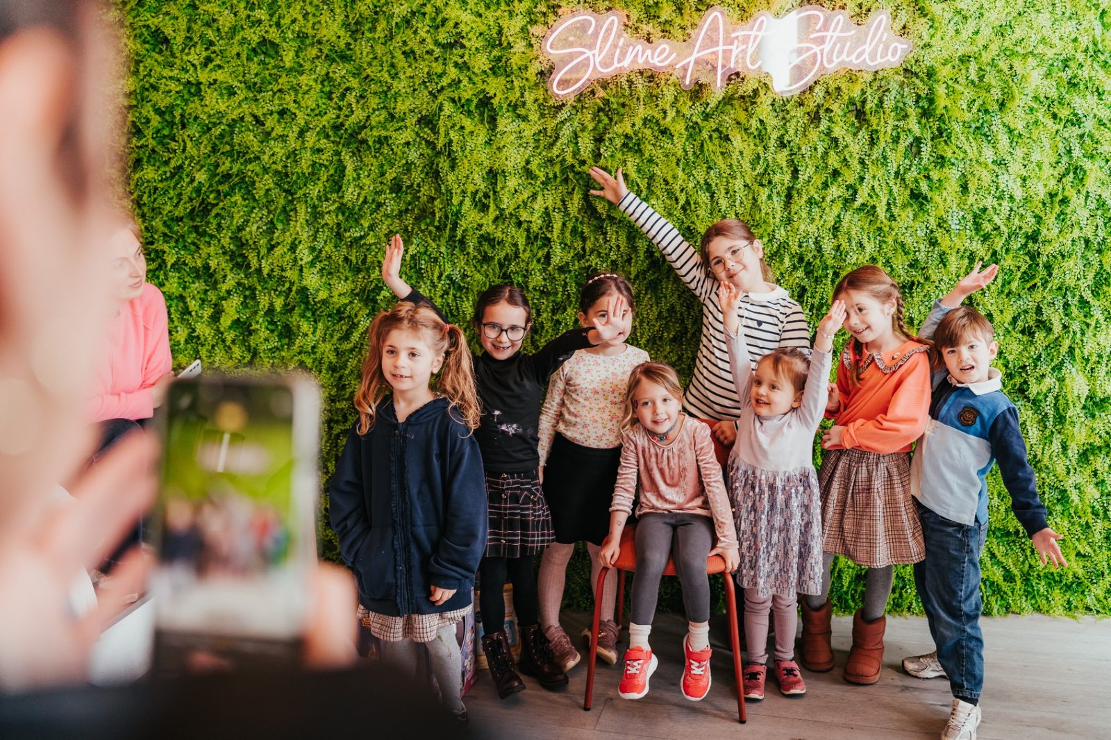 Atelier slime à Waterloo — groupe d'enfants souriant pour une photo souvenir devant le mur végétal et l'enseigne néon Slime Art Studio